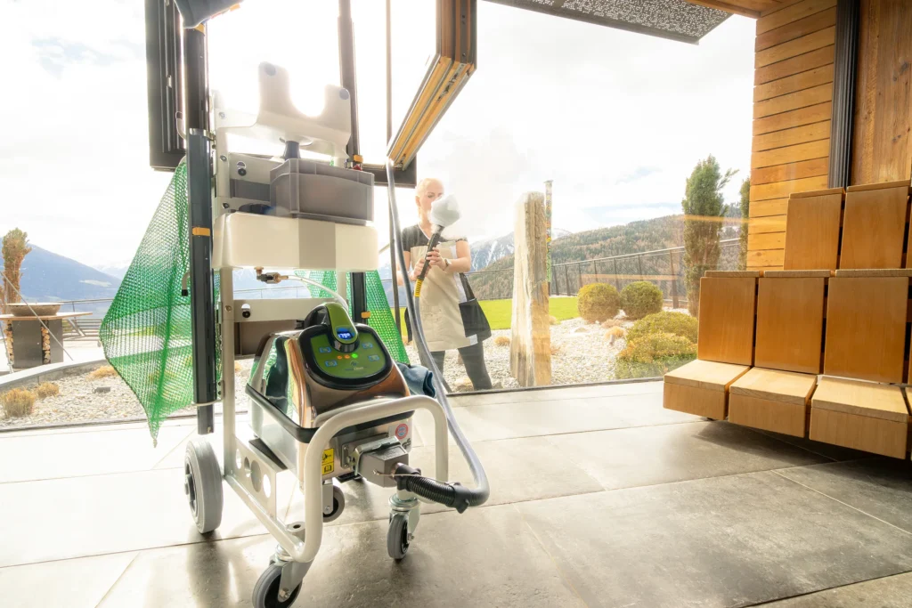 MediCleanTEC Dampfreiniger mit grünem Griff auf einem Gerätewagen mit Zubehör im Zentrum des Bildes, in einer Sauna. Dahinter große Fenster mit Blick nach draußen, wo eine Dame die Fensterfläche von außen mit der Dreiecksbürste inklusive weißem Baumwolltuch reinigt. Der lange Schlauch wird dabei von innen durch das geöffnete Fenster nach draußen gereicht.